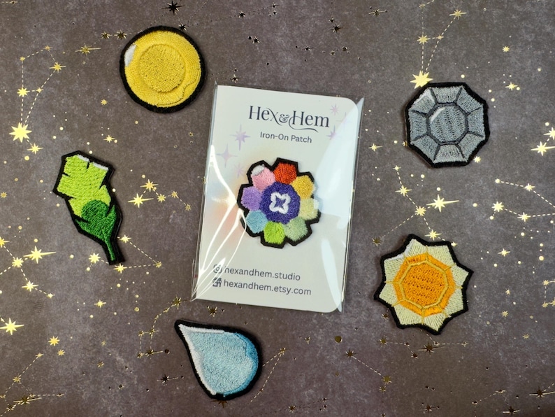 Puede incluir: Un surtido de parches termoadhesivos, que incluyen un c&iacute;rculo amarillo, una pluma verde, una flor multicolor, una forma geom&eacute;trica gris, un sol amarillo y una l&aacute;grima azul. Los parches est&aacute;n sobre un fondo oscuro con patrones de estrellas doradas.