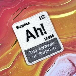 May include: A white embroidered patch with the text "Surprise" at the top and "Ah!" in large black letters. Below, it reads "The Element of Surprise." The patch has a black border and the number "117" is visible.