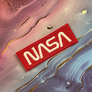 May include: A rectangular red embroidered patch with the white letters "NASA". The patch is a rectangle with rounded corners. The background is a colorful mix of pink, blue, and gold.