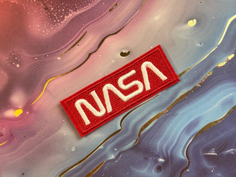 May include: A rectangular red embroidered patch with the white letters "NASA". The patch is a rectangle with rounded corners. The background is a colorful mix of pink, blue, and gold.