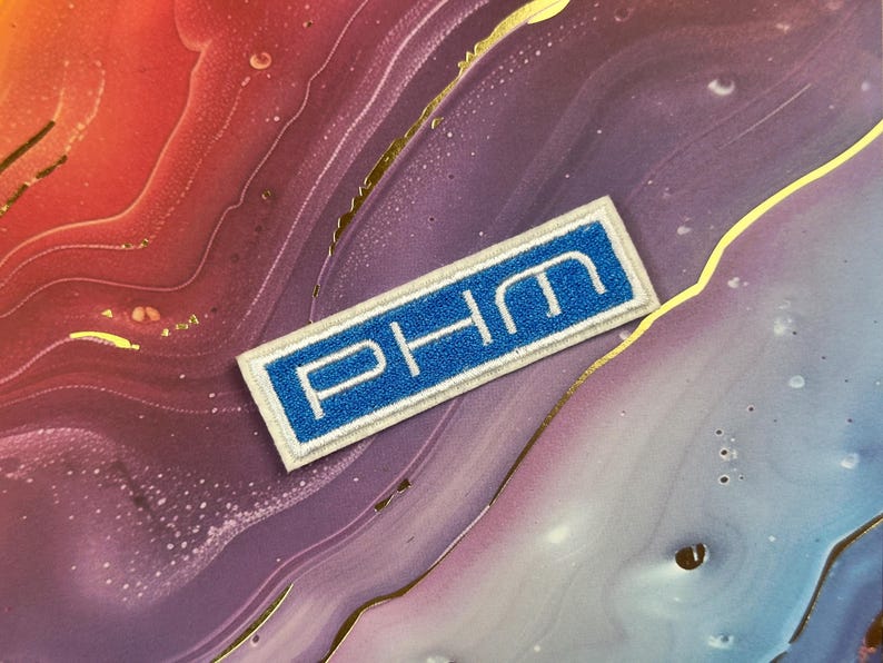 May include: A rectangular patch with the letters "PHM" in white, set against a blue background. The patch has a white border and is displayed on a colorful, marbled surface with gold accents.