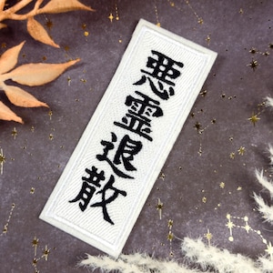 May include: A rectangular white patch with black Japanese characters embroidered on it. The patch is bordered with white stitching. The background is a dark gray surface with gold star-shaped confetti and dried plants.