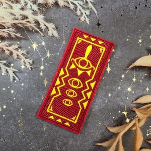 Puede incluir: Un parche rectangular rojo con un diseño de ojo amarillo. El parche presenta una superficie texturizada y un borde amarillo. El diseño incluye un ojo grande en la parte superior y tres ojos más pequeños debajo. Probablemente sea un parche para planchar o coser.