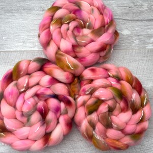 May include: Three braided bundles of soft roving wool fiber, each featuring a variegated color palette of bright pink, deep magenta, and earthy olive green tones, arranged in a triangular formation with a thick, textured, and coiled appearance.