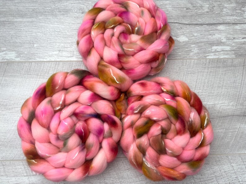 May include: Three braided bundles of soft roving wool fiber, each featuring a variegated color palette of bright pink, deep magenta, and earthy olive green tones, arranged in a triangular formation with a thick, textured, and coiled appearance.