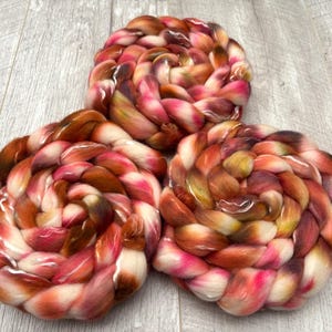 May include: Three skeins of hand-dyed yarn in a spiral arrangement. The yarn features a blend of colors, including shades of brown, pink, and white. The texture of the yarn appears soft and fluffy, ideal for knitting or crocheting projects.