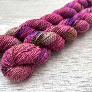 May include: Close-up of several skeins of hand-dyed yarn in varying shades of pink, purple, and brown. The yarn is arranged diagonally across a light-coloured wooden surface, showcasing the vibrant colours and textures of the fibre. Ideal for knitting and crochet projects.