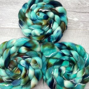 Puede incluir: Tres ovillos circulares de lana peinada con una paleta de colores variados en tonos turquesa, azul cerceta, azul marino profundo y marr&oacute;n tierra. Las fibras suaves y gruesas est&aacute;n trenzadas en espirales densas con una textura lisa y brillante.