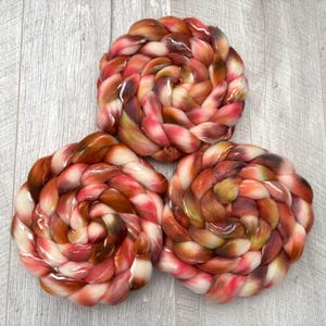 May include: Three skeins of hand-dyed yarn in a spiral arrangement. The yarn features a blend of colors, including shades of red, brown, and cream. The yarn is arranged on a light gray wooden surface.