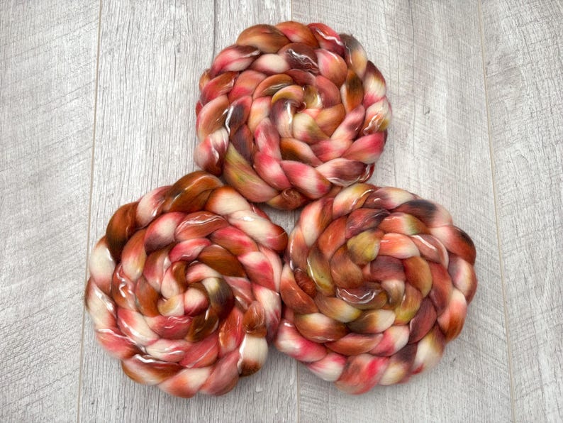 May include: Three skeins of hand-dyed yarn in a spiral arrangement. The yarn features a blend of colors, including shades of red, brown, and cream. The yarn is arranged on a light gray wooden surface.