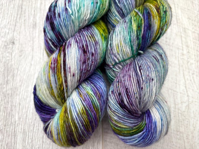 May include: Two skeins of hand-dyed yarn in a vibrant mix of colors. The yarn features shades of purple, blue, green, and yellow, creating a speckled effect. The yarn is wound into a soft, textured bundle, perfect for knitting or crocheting projects.