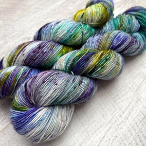 May include: A skein of hand-dyed yarn in shades of blue, green, purple, and yellow. The yarn is arranged in a loose, flowing shape, with the colors blending together to create a variegated effect. The yarn appears soft and textured.