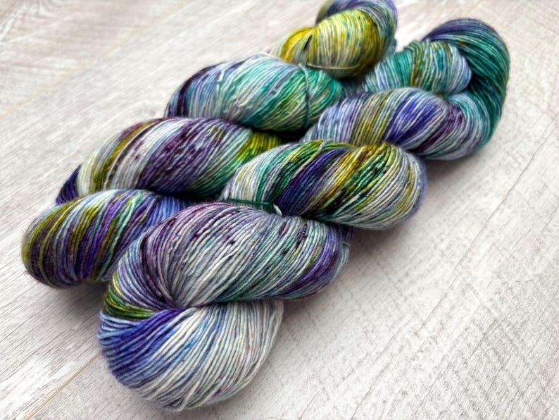May include: A skein of hand-dyed yarn in shades of blue, green, purple, and yellow. The yarn is arranged in a loose, flowing shape, with the colors blending together to create a variegated effect. The yarn appears soft and textured.