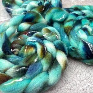 Puede incluir: Dos gruesas trenzas de lana peinada suave. La fibra presenta una paleta de colores variados de turquesa vibrante, azul marino profundo y tonos marrones terrosos, mezclados para crear una apariencia multicolor y texturizada.