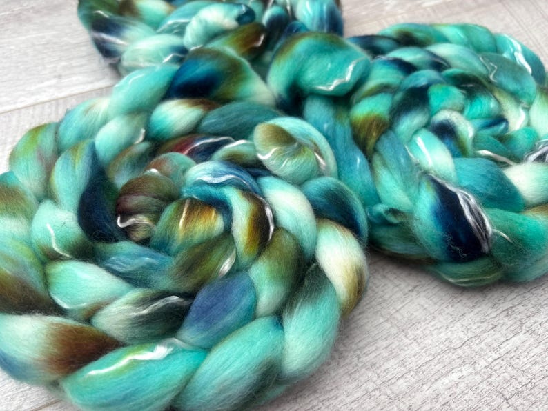 Puede incluir: Dos gruesas trenzas de lana peinada suave. La fibra presenta una paleta de colores variados de turquesa vibrante, azul marino profundo y tonos marrones terrosos, mezclados para crear una apariencia multicolor y texturizada.