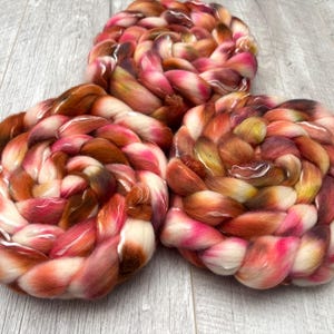May include: Three skeins of hand-dyed yarn in a braided, circular shape. The yarn features a blend of colors, including shades of pink, orange, brown, and white. The yarn is arranged on a light gray wooden surface.