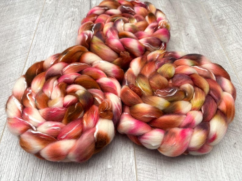 May include: Three skeins of hand-dyed yarn in a braided, circular shape. The yarn features a blend of colors, including shades of pink, orange, brown, and white. The yarn is arranged on a light gray wooden surface.