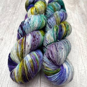 May include: Two skeins of hand-dyed yarn in a vibrant array of colors. The yarn features a blend of blues, greens, purples, and yellows, creating a speckled effect. The yarn is arranged on a light-colored wooden surface.