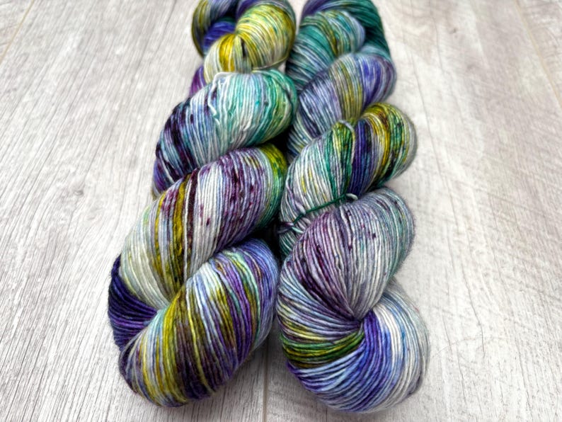 May include: Two skeins of hand-dyed yarn in a vibrant array of colors. The yarn features a blend of blues, greens, purples, and yellows, creating a speckled effect. The yarn is arranged on a light-colored wooden surface.
