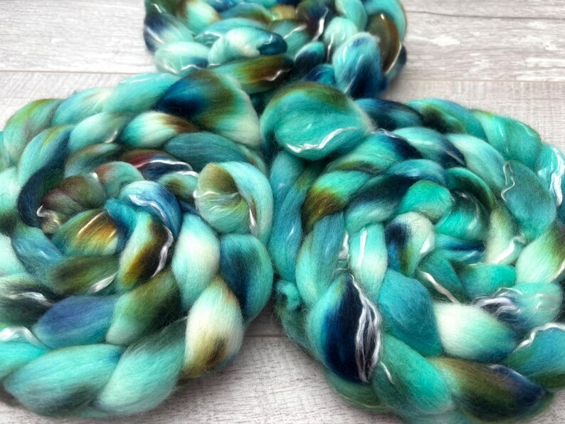 Puede incluir: Tres gruesas trenzas de lana cardada suave. El hilo presenta una paleta de colores variados de turquesa brillante, azul marino profundo y verde oliva terroso, mezclados en un patr&oacute;n de espiral continuo, suave y de textura esponjosa.