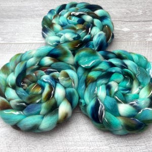 Puede incluir: Tres madejas trenzadas de lana cardada con una paleta de colores variados de turquesa vibrante, verde azulado profundo, azul marino oscuro y tonos marrones terrosos, dispuestas en forma triangular con una apariencia suave y fibrosa.