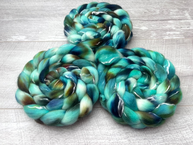 Puede incluir: Tres madejas trenzadas de lana cardada con una paleta de colores variados de turquesa vibrante, verde azulado profundo, azul marino oscuro y tonos marrones terrosos, dispuestas en forma triangular con una apariencia suave y fibrosa.