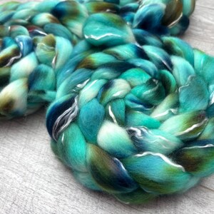 Puede incluir: Una mecha de lana gruesa y trenzada con una paleta de colores variados de turquesa vibrante, verde bosque profundo y azul marino oscuro. La textura suave y esponjosa de la fibra sin hilar est&aacute; enrollada en forma circular con hebras visibles.