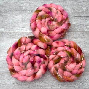 May include: Three circular braids of thick, soft yarn arranged in a triangular formation. The yarn features a variegated color palette of bright pink, deep magenta, and earthy brown tones, creating a marbled effect throughout the braided texture.