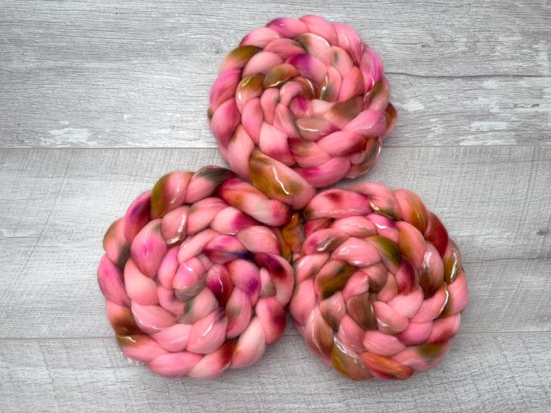 May include: Three circular braids of thick, soft yarn arranged in a triangular formation. The yarn features a variegated color palette of bright pink, deep magenta, and earthy brown tones, creating a marbled effect throughout the braided texture.
