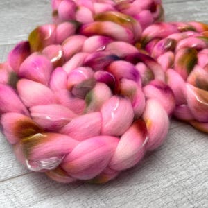 May include: A thick, braided roving of soft fiber in vibrant shades of bright pink, magenta, and deep orange-brown. The material is coiled into a circular shape, showcasing a smooth, voluminous texture with blended color transitions throughout the strands.