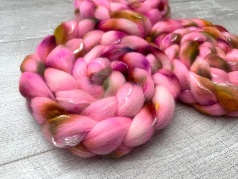 May include: A thick, braided roving of soft fiber in vibrant shades of bright pink, magenta, and deep orange-brown. The material is coiled into a circular shape, showcasing a smooth, voluminous texture with blended color transitions throughout the strands.