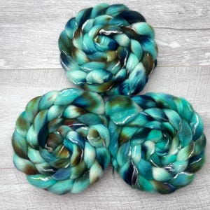 Puede incluir: Tres trenzas circulares de fibra de lana peinada con una paleta de colores variados en verde azulado, turquesa, azul profundo y marr&oacute;n oscuro. Las fibras est&aacute;n retorcidas en formas espirales apretadas, mostrando una textura suave.