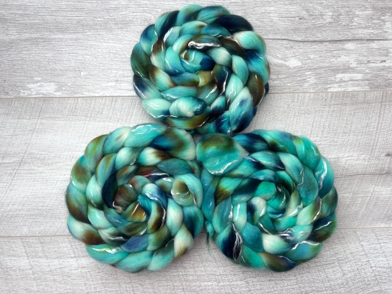 Puede incluir: Tres trenzas circulares de fibra de lana peinada con una paleta de colores variados en verde azulado, turquesa, azul profundo y marr&oacute;n oscuro. Las fibras est&aacute;n retorcidas en formas espirales apretadas, mostrando una textura suave.