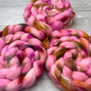May include: Three circular coils of thick, braided roving wool yarn. The fiber features a variegated color palette of bright pink, deep magenta, and earthy olive green tones, arranged in a dense, soft, and textured spiral pattern.