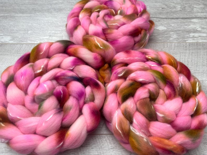 May include: Three circular coils of thick, braided roving wool yarn. The fiber features a variegated color palette of bright pink, deep magenta, and earthy olive green tones, arranged in a dense, soft, and textured spiral pattern.