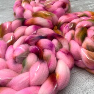 May include: Three thick, braided coils of soft roving wool. The fiber features a variegated color palette dominated by bright pink, with blended accents of olive green, deep brown, and hints of dark purple throughout the textured, fluffy strands.
