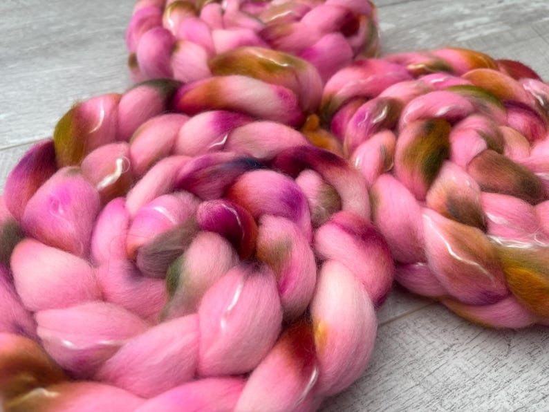 May include: Three thick, braided coils of soft roving wool. The fiber features a variegated color palette dominated by bright pink, with blended accents of olive green, deep brown, and hints of dark purple throughout the textured, fluffy strands.