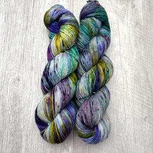 May include: Two skeins of hand-dyed yarn in a vibrant array of colors. The yarn features a blend of blues, greens, purples, and yellows, creating a speckled and variegated effect. The yarn is arranged on a light wood-grain surface.