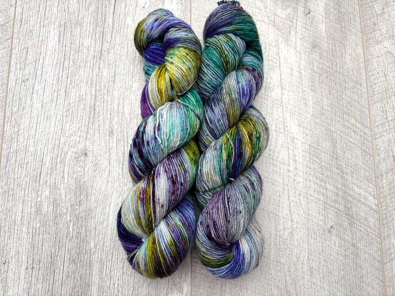 May include: Two skeins of hand-dyed yarn in a vibrant array of colors. The yarn features a blend of blues, greens, purples, and yellows, creating a speckled and variegated effect. The yarn is arranged on a light wood-grain surface.