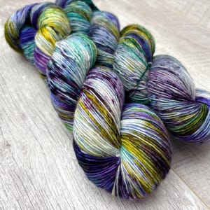 May include: A skein of hand-dyed yarn in shades of blue, purple, green, and yellow. The yarn is arranged in a loose bundle, with the colors blending together to create a variegated effect. The yarn is on a light wood surface.