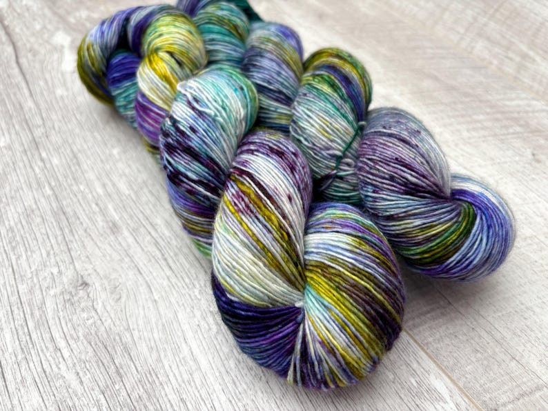 May include: A skein of hand-dyed yarn in shades of blue, purple, green, and yellow. The yarn is arranged in a loose bundle, with the colors blending together to create a variegated effect. The yarn is on a light wood surface.