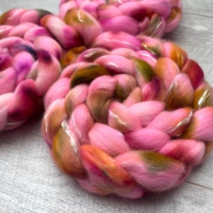 May include: Three thick, braided coils of soft roving wool fiber. The wool features a variegated color palette of bright pink, deep magenta, warm orange, and earthy olive green tones, blended together in a continuous, soft, and fluffy texture.