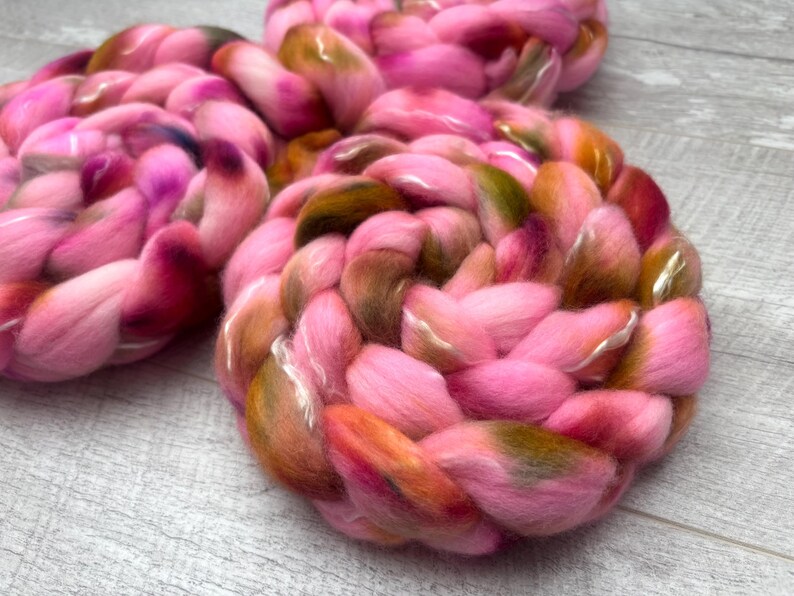 May include: Three thick, braided coils of soft roving wool fiber. The wool features a variegated color palette of bright pink, deep magenta, warm orange, and earthy olive green tones, blended together in a continuous, soft, and fluffy texture.