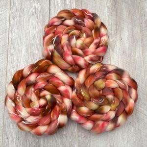 May include: Three swirled bundles of colorful yarn in shades of red, brown, and cream. The yarn is arranged in a spiral pattern, creating a visually appealing texture. The background is a light gray wooden surface.