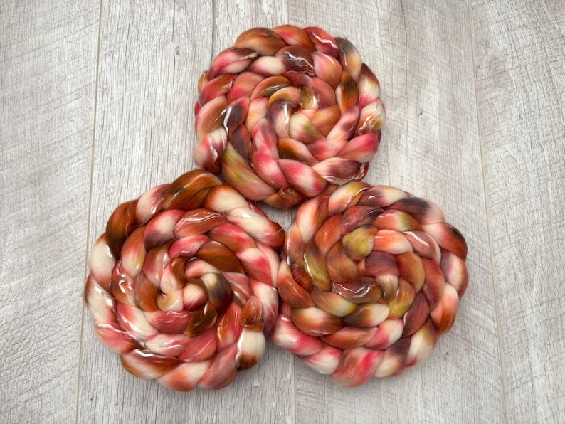 May include: Three swirled bundles of colorful yarn in shades of red, brown, and cream. The yarn is arranged in a spiral pattern, creating a visually appealing texture. The background is a light gray wooden surface.