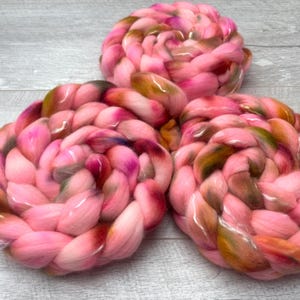 May include: Three circular coils of thick, braided roving wool yarn. The fiber features a variegated color palette of bright pink, soft rose, deep burgundy, and earthy olive green tones, twisted together to create a textured, voluminous appearance.