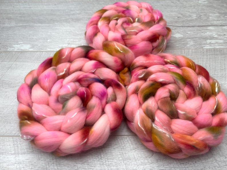 May include: Three circular coils of thick, braided roving wool yarn. The fiber features a variegated color palette of bright pink, soft rose, deep burgundy, and earthy olive green tones, twisted together to create a textured, voluminous appearance.