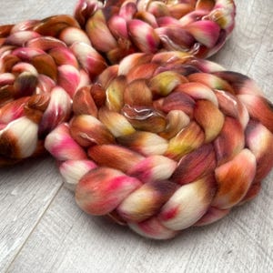 May include: Three skeins of hand-dyed yarn in a braided pattern. The yarn features a blend of colors, including white, pink, brown, and orange. The yarn is arranged on a light gray wooden surface.