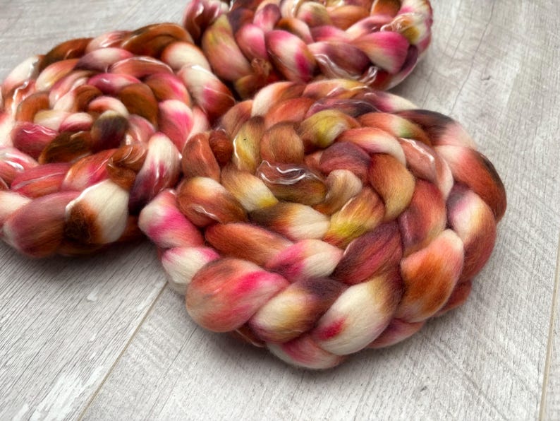 May include: Three skeins of hand-dyed yarn in a braided pattern. The yarn features a blend of colors, including white, pink, brown, and orange. The yarn is arranged on a light gray wooden surface.