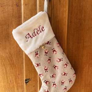 Könnte beinhalten: Ein beiger Weihnachtssocken mit einem weißen Kunstfell-Manschetten. Der Socken ist mit einem sich wiederholenden Muster aus roten und schwarzen Weihnachtsmann-Figuren verziert. Die Manschette ist mit dem Namen "Adèle" in roter Farbe bestickt.